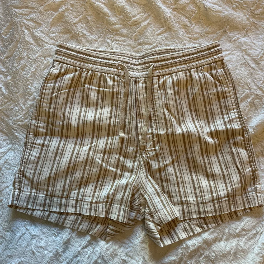 Säljer ett par bekväma och stilrena randiga shorts från H&M i storlek XS. De är i en ljus beige färg med bruna ränder och har en elastisk midja med dragsko för perfekt passform. Shortsen har en bakficka och är gjorda i ett lätt och luftigt material, perfekt för varma sommardagar.. Shorts.