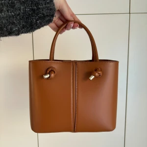 Handväska från Zara - En brun handväska från Zara. Superfin men ej använd. Finns ett långt band till som man kan ta av om man vill. Fack i mitten med dragkedja och stängning med knapp upptill.  Mått: 23x18x10