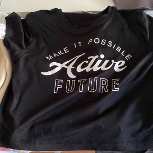 Svart t-shirt från Kappahl - Säljer en svart t-shirt från Kappahl med texten 'Make it Possible Active Future' tryckt i vitt på framsidan. T-shirten är kortärmad och perfekt för en aktiv vardag. Passar bra till både träning och fritid!