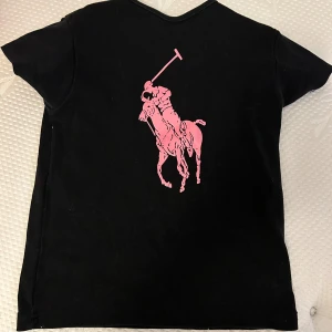 Svart t-shirt från Ralph Lauren med rosa tryck - Säljer en snygg svart t-shirt från Ralph Lauren med ett stort rosa polospelartryck på framsidan . T-shirten är kortärmad och har en klassisk passform. Sitter son baby shirt.
