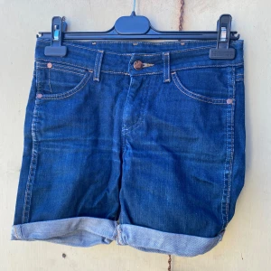 Jeansshorts från Wrangler - Säljer ett par snygga jeansshorts från Wrangler. De är mörkblå med klassisk femficksdesign och har en uppvikt kant nedtill. Perfekta för sommaren och har en skön passform. Materialet är 98% bomull och 2% elastan vilket ger lite stretch.