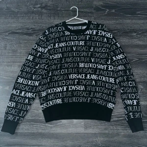 Svart tröja från Versace Jeans Couture - Säljer en snygg svart tröja från Versace Jeans Couture. Den har ett coolt mönster med märkesnamnet i vitt över hela tröjan. Perfekt för att ge din outfit en lyxig touch. Tröjan är långärmad och har ribbade muddar vid ärmslut och nederkant. Passar perfekt för både vardag och fest!