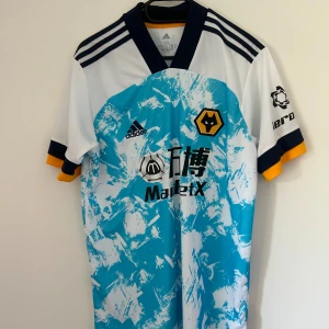 Adidas fotbollströja med blått mönster - Wolves borta fotbollströja från säsong 2020/21. Wolves hamnade på plats 13 i premier league under den säsongen. Tröjan är fel fri.
