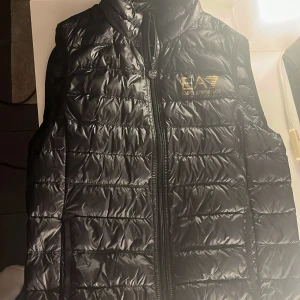 Svart Armani dunväst - Säljer nu min emporio Armani dunväst då den är för liten. Väldigt fint skick, köpt på Zalando. Nypris 1999kr. Tillkommer en bag som man kan fästa på insidan av västen (bild 3). 2 fickor på utsidan och en på vänster insida.