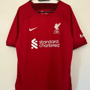 Säljer en röd Liverpool fotbollströja från Nike. Tröjan har korta ärmar och är i Dri-FIT material för bra andningsförmåga. På framsidan finns Liverpool FC:s emblem och sponsorlogga, medan baksidan har namnet 'VIRGIL' och nummer 4. Tröjan är replika.