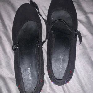 Svarta ballerinaskor med färgglada pärlor - Säljer ett par svarta ballerinaskor från Graceland. De är dekorerade med färgglada pärlor som ger en festlig touch. Skorna har en rem över foten för extra passform och komfort. Perfekta för både vardag och fest!