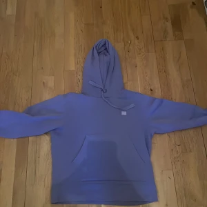 Lila hoodie från Acne Studios - Säljer en superfin lila hoodie från Acne Studios. Den är i nyskick och har en klassisk design med en stor ficka fram och justerbar huva. Perfekt för både höst och vår när man vill vara både bekväm och stilren. Materialet känns mjukt och skönt mot huden.