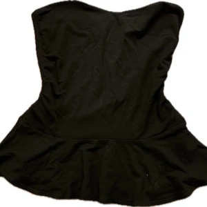 Svart axelbandslös topp - Snygg svart axelbandslös topp med en figurnära passform och volangdetalj vid nederkanten. Perfekt för en kväll ute eller en speciell tillställning.