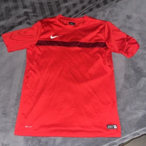 Röd fotbollströja från Nike - Säljer en röd Nike fotbollströja med korta ärmar och Dri-FIT material för optimal komfort. Tröjan har en mörkröd rand över bröstet och en vit Nike-logga. Perfekt för träning eller match! Väl använd men har inga defekter