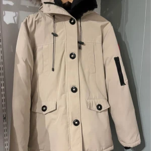 Beige parkajacka från Canada Goose - Säljer en snygg beige parkajacka från Canada Goose med pälsdetaljer på huvan. Jackan har långa ärmar, dragkedja och knappar framtill samt en ikonisk logga på ärmen. 
