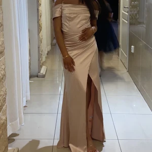Champagnefärgad långklänning - Elegant champagnefärgad långklänning med offshoulder-design och en stilfull slits framtill. Perfekt för en festlig kväll.