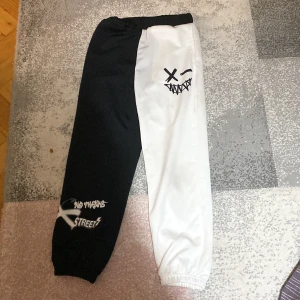 Svartvita byxor med grafiskt tryck - Unika byxor i svart och vitt med ett coolt grafiskt tryck. Ena benet är svart med texten 'No Friends Street' och det andra benet är vitt med ett ansikte som har korsade ögon och ett brett leende. Perfekta för en streetwear-look.