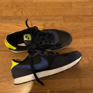 Nike sneakers med blå och gula detaljer - Snygga Nike sneakers i svart med blå swoosh och gula detaljer på hälen. Skorna har en klassisk design med snörning och en bekväm sula. Perfekta för en sportig look. perfekt skick. 