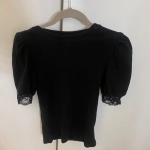 Svart topp med spetsdetaljer från Name It - Säljer en svart topp från Name It med korta puffärmar och fina spetsdetaljer vid ärmsluten. Står att den passar åldrar 11-12 år. Men skulle mer säga 13-14 eller som en S