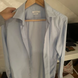 Ljusblå skjorta från J.Lindeberg - Snygg ljusblå skjorta från J.Lindeberg i slim fit. Perfekt för en stilren look med klassiska knappar framtill. Skjortan har långa ärmar och är tillverkad i ett mjukt material som ger en bekväm känsla.