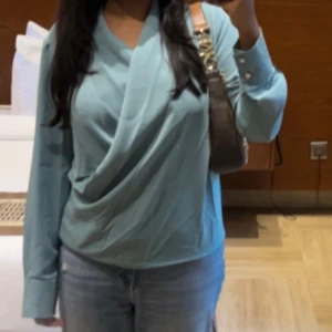 Turkos blus - Snygg turkos omlott-aktig blus med långa ärmar och elegant drapering framtill. Perfekt för en stilren look.