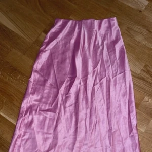 Rosa långkjol - Snygg rosa långkjol i ett mjukt och glansigt material. Perfekt för en färgglad look. Kjolen har en enkel och elegant design som gör den lätt att matcha med olika toppar.