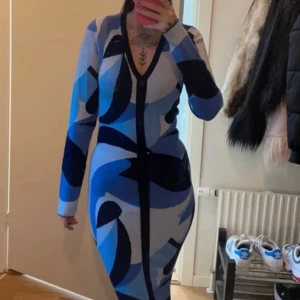 Mönstrad långklänning i blå nyanser - Snygg långklänning med ett unikt mönster i olika blå nyanser. Klänningen har en v-ringad design och långa ärmar, perfekt för en stilfull look. Den är figurnära och framhäver silhuetten på ett elegant sätt.