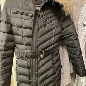 Svart dunjacka med päls från Hollies - Säljer en snygg svart dunjacka från Hollies med pälsdetalj på huvan. Jackan har dragkedja och bälte i midjan för en figursydd look. Insidan är fodrad med ett coolt leopardmönster. Perfekt för kalla dagar!