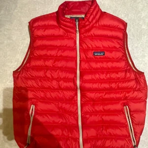 dunväst Patagonia - Säljer en snygg röd dunväst från Patagonia med dragkedja framtill. Västen har en quiltad design och två praktiska fickor med dragkedja. Perfekt för kyligare dagar och ger en sportig look.