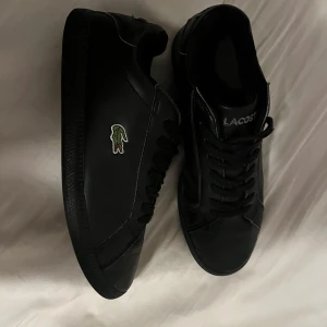 Svarta sneakers från Lacoste - Snygga svarta sneakers från Lacoste med klassisk krokodillogga på sidan. Skorna har en stilren design med snörning och är tillverkade i skinn. Perfekta för en casual look. Perfekt skick förutom snörena lite skadade längst upp, går att fixa.