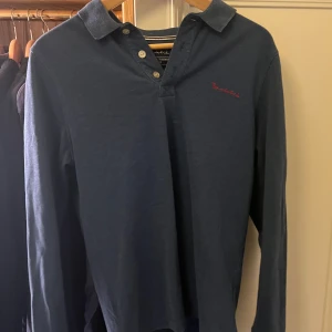 Mörkblå pikétröja från Brooks Brothers - Snygg mörkblå pikétröja från Brooks Brothers med broderad logga i rött på bröstet. Tröjan har långa ärmar och klassisk krage med knappar. Perfekt för en stilren look.