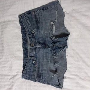 Blå lågmidjade jeansshorts - Säljer ett par blå jeansshorts med råa kanter och slitna detaljer. Shortsen har en knapp och dragkedja framtill samt två bakfickor med dekorativa sömmar. Perfekta för en avslappnad stil. Lappen är borta så vet tyvärr inte vilket märke. Jag uppskattar storleken att vara 36