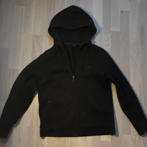 Svart hoodie från Hugo Boss - Säljer en stilren svart hoodie från Hugo Boss med dragkedja och justerbar huva. Perfekt för en avslappnad look. Diskret logga på bröstet och ribbade muddar vid ärmslut och nederkant.