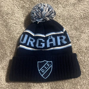 Blå mössa med DIF-logga - Snygg blå mössa med DIF-logga framtill. Mössan har en stor tofs på toppen och texten 'DJURGÅRDEN' i ljusblått. Perfekt för kalla dagar och visar ditt stöd för laget. Nyskick. Helt slutsåld i både butik och på nätet, nypris 299. Kan även tänka mig byta mot en rosa 