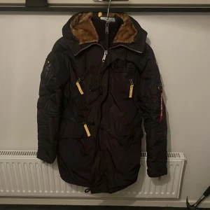Svart vinterjacka från Alpha Industries - Säljer en svart vinterjacka från Alpha Industries med gul insida och detaljer. Jackan har en pälsfodrad huva och flera fickor med dragkedjor. Perfekt för kalla dagar med sin quiltade insida och långa ärmar. Huvan och ärmsluten har gula dragkedjor för extra stil. 
