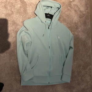 Ljusblå hoodie från Polo Ralph Lauren - Säljer en stilren ljusblå hoodie från Polo Ralph Lauren. Tröjan har en dragkedja framtill och en liten broderad logga på bröstet. Perfekt för en avslappnad stil. 