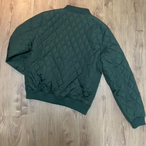 Grön quiltad bomberjacka från Urban Classics - Säljer en snygg grön quiltad bomberjacka från Urban Classics. Jackan har en dragkedja framtill och ribbade muddar vid ärmslut och nederkant. Perfekt för en stilren look.