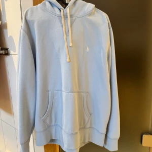 Ljusblå hoodie från Ralph Lauren - Säljer en stilren ljusblå hoodie från Ralph Lauren med klassisk känguruficka och justerbar huva med snören. Den har en liten broderad logga på bröstet. Perfekt för en avslappnad stil.