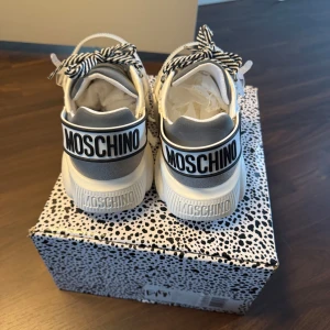 Vita sneakers från Moschino - Snygga vita sneakers från Moschino med svart text på hälen. Skorna har en unik sula med en björnformad design och randiga snören. De är tillverkade i syntetmaterial med transparenta detaljer och en modern look. Aldrig använda köpta för 3500  