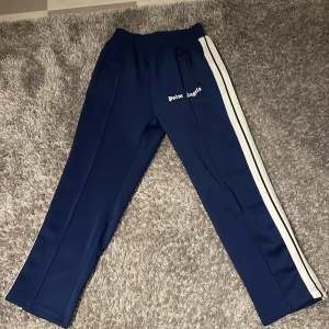 Blå Palm Angels byxor - Snygga blå träningsbyxor från Palm Angels med vita ränder längs sidorna. Byxorna har en elastisk midja och en avslappnad passform. Perfekta för en sportig look.