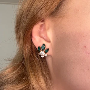 Clipps örhängen med gröna stenar  - Eleganta clipps örhängen med gröna och klara stenar i en blommig design. Perfekta för att ge en touch av glamour till din look.