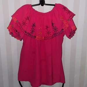 Rosa singoalla topp  med broderi st.162-164 - Charmig rosa singoalla topp med vackra broderade detaljer i blått och gult. Toppen har en offshoulder-design med volang runt axlarna, vilket ger en söt och lekfull look. Perfekt för somriga dagar och speciella tillfällen.
