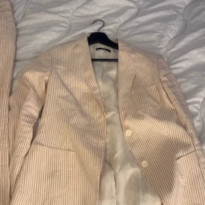 Randig kostym set från Hugo boss - Snygg randig kostym i beige och vit med kavaj och byxor. Tillverkad i en lyxig blandning av linne och siden. Kavajen har knappar framtill och byxorna har en klassisk passform med bälte. Perfekt för en stilren look. Kostym byxorna är låidjade och inte alls töjbara helt nya oxå!!! Byxorna är i storlek 40 och kavajen i 38