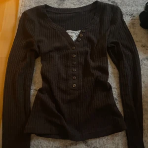Brun ribbad tröja med knappar - Säljer en brun ribbad tröja med långa ärmar och knappar framtill. Tröjan har en v-ringad design med spetsdetalj vid halsen. 