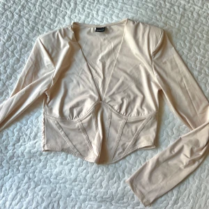 Beige långärmad korsettopp - Snygg beige korsettopp från Gina med långa ärmar och en djup v-ringning. Toppen är croppad och har en stilren design som passar perfekt för en trendig look.