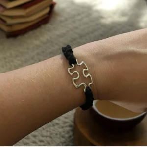 Svart armband med pusselbit i silver - Snyggt armband med en unik pusselbitsdetalj i silver. Bandet är svart och knuten med makramé, vilket ger en stilren och modern look. Perfekt för att ge en personlig touch till din stil.