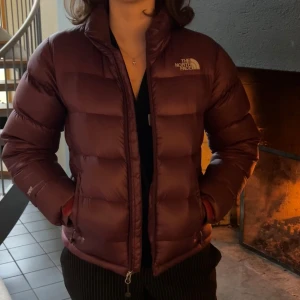 Vinröd dunjacka från The North Face - Säljer en snygg vinröd dunjacka från The North Face. Jackan har en dragkedja framtill och är perfekt för kyliga dagar. Den är långärmad och har en klassisk pufferdesign som håller dig varm och stilren. Fin vinter / vårjacka från the north face i storlek S. Säljer då den tyvärr är förliten för mig. Finns ett litet hål vid ena fickan