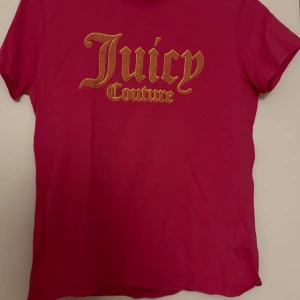 Rosa t-shirt från Juicy Couture - Säljer en snygg rosa t-shirt från Juicy Couture med deras ikoniska logga i guld på framsidan. T-shirten är kortärmad och tillverkad i 100% bomull, vilket gör den mjuk och bekväm att bära. Perfekt för en avslappnad stil. Storleken motsvarar typ S
