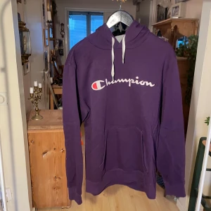 Lila hoodie från Champion - Säljer en snygg lila hoodie från Champion med vit logga på bröstet. Tröjan har en klassisk känguruficka och justerbar huva med vita snören. Perfekt för en avslappnad stil.