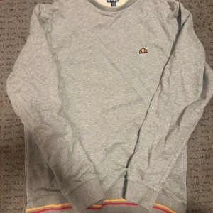 Grå tröja från Ellesse - Säljer en grå tröja från Ellesse med färgglada ränder i gult och rött vid nederkanten. Tröjan har långa ärmar och en liten logotyp på bröstet. Perfekt för en avslappnad stil. Helt ny. Bara avhämtning i Göteborg eller Mölndal. Ingen frakt!