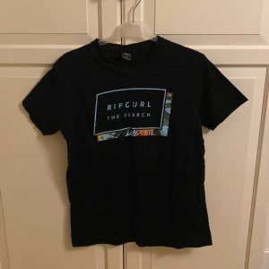 Svart t-shirt från Rip Curl - Snygg svart t-shirt från Rip Curl med tryck 'The Search' och färgglada detaljer. Perfekt för en avslappnad stil. Kortärmad och bekväm passform. I fint skick. Storlek S.