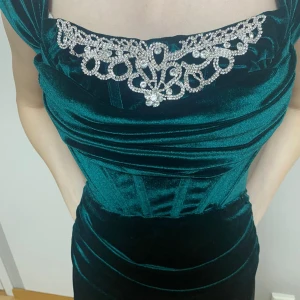 Grön sammetsklänning med strassdetaljer - Elegant grön långklänning i sammet med vackra strassdetaljer vid halsringningen. Klänningen har en figurnära passform och är perfekt för festliga tillfällen. Den djupa gröna färgen ger en lyxig känsla.