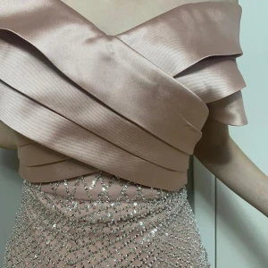 Champagnefärgad långklänning med paljetter - Elegant champagnefärgad långklänning med offshoulder-design och glittrande paljettdetaljer. Klänningen har en omlott-topp och ett vackert fall med ett nätmönster av paljetter som ger en lyxig känsla.