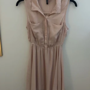 Ljusrosa ärmlös klänning - Säljer en söt ljusrosa ärmlös klänning med krage och knappar framtill. Klänningen har en elastisk midja som ger en fin passform och en luftig kjol. Perfekt för varma dagar!