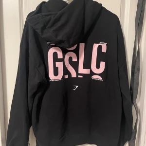Gymshark hoodie - Superskön Gymshark hoodie! Strl M och har en oversized fit🩷 ljuset på bilderna är lite halvskevt men den är kolsvart och har rosa och vit text på trycket🥰köpt för 499 och ganska sparsamt använd! Säljer för att jag har för många